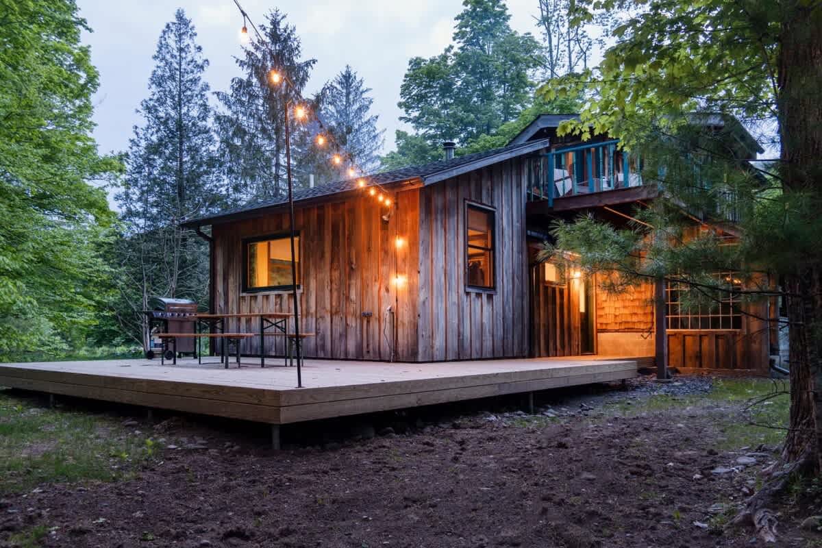 The cabin at 1392 Route 214 at dusk, glowing under string lights with a wood deck and dining table