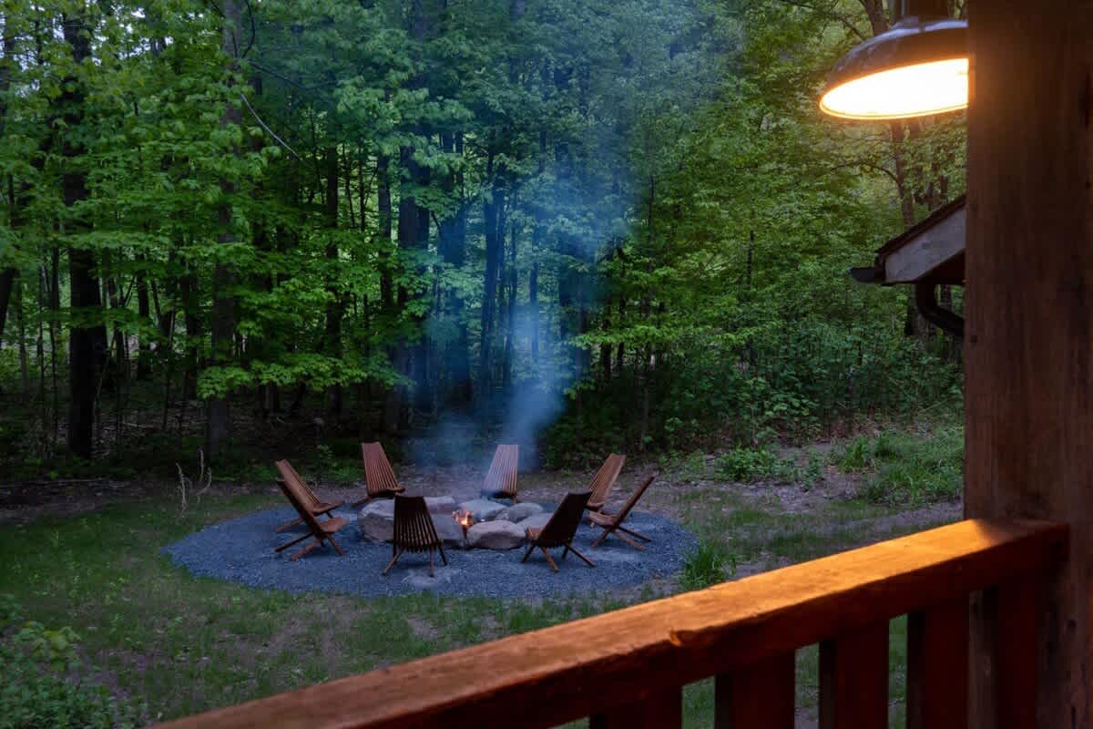 A stone fire pit ringed by wooden chairs in the woods behind 1392 Route 214 at dusk