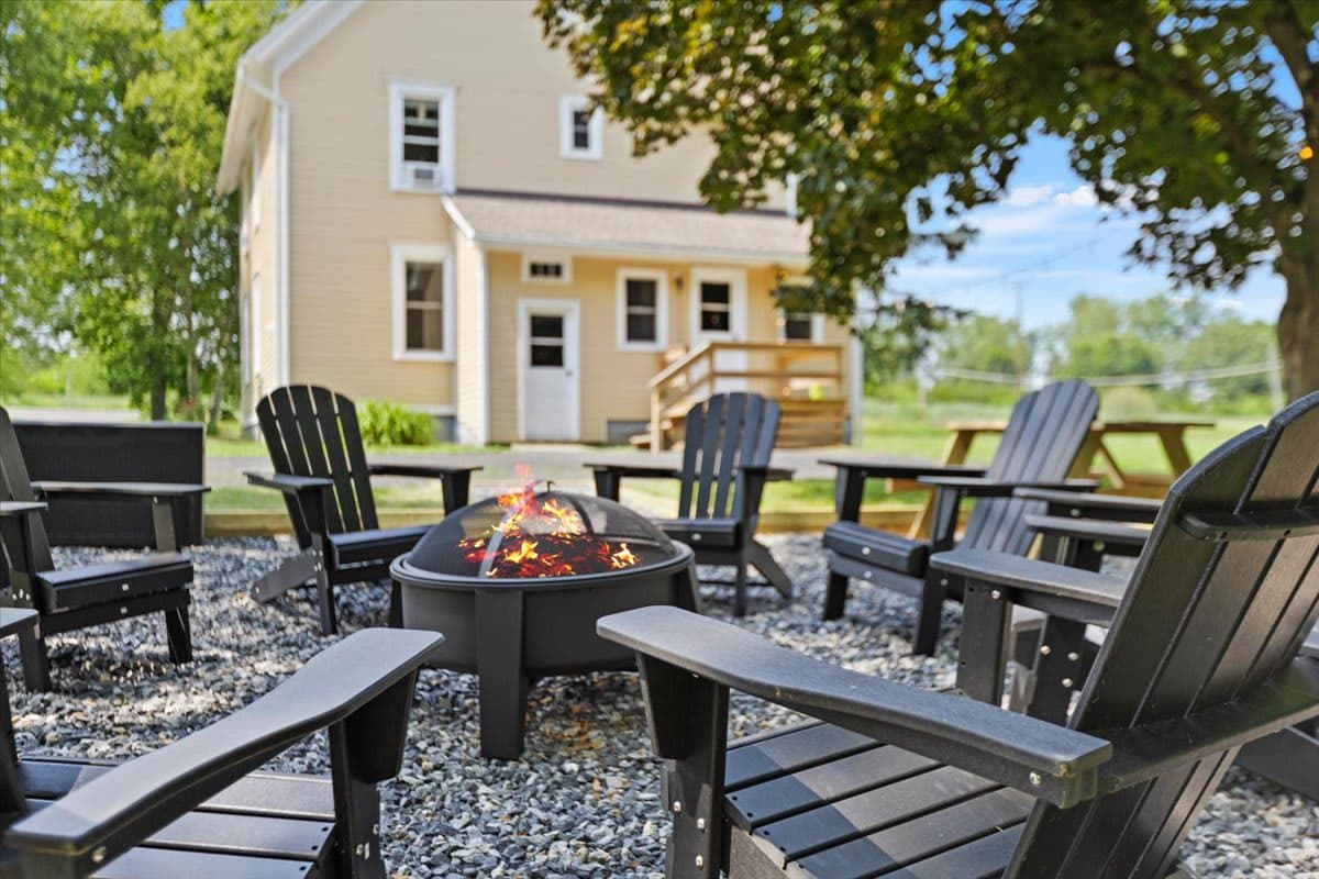 Black Adirondack chairs around a lit fire pit on the gravel patio behind the yellow farmhouse